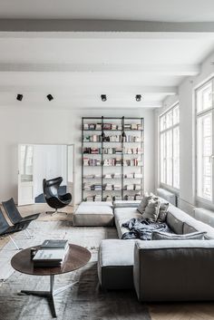 a living room filled with lots of furniture next to two large windows and bookshelves