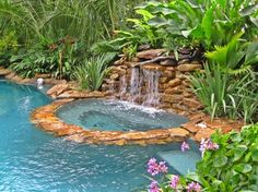 an outdoor swimming pool surrounded by greenery and rocks with a waterfall in the middle