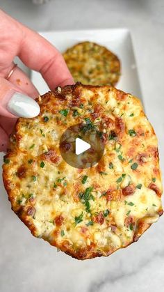 a person holding up a small pizza with cheese and toppings on it, in front of another food item