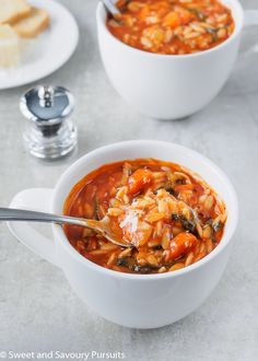 two bowls of pasta soup on a table with bread and silverware in the background