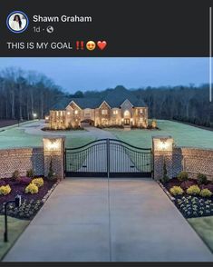 a driveway leading to a large home with lights on the front gate and landscaping around it