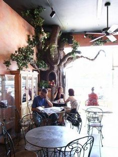 people are sitting at tables in an outdoor cafe with a tree growing on the wall