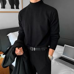 a man standing in front of a bed wearing a black turtle neck sweater and pants