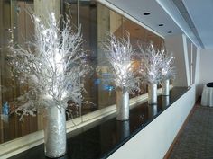 three silver vases with white branches in them on a black counter next to a mirror