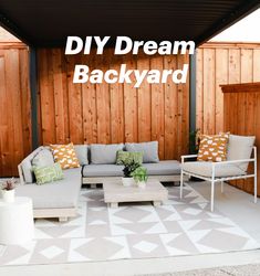 an outdoor living area with couches, tables and pillows on the floor in front of a wooden fence