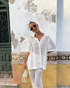 a woman standing on the street wearing white clothing and holding a handbag in her right hand