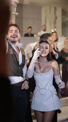 a man in a tuxedo holding a wine glass next to a woman in a short white dress