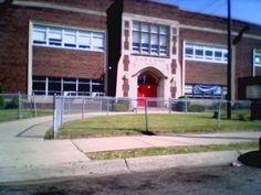 an old school building with a red door and fence around the front yard is shown