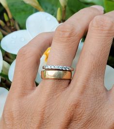 a person's hand with a gold ring on it and flowers in the background