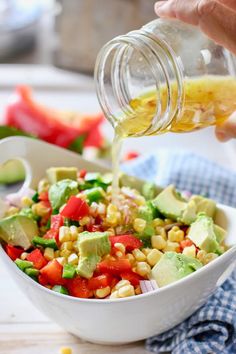 a person pouring dressing into a bowl filled with corn, avocado and tomatoes