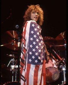 a woman standing in front of a drum set with an american flag draped over her