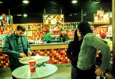 two people standing in front of a counter at a fast food restaurant, one person is cutting pizza