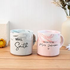 two coffee mugs sitting next to each other on top of a wooden table with flowers in the background