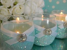 three clear glass candles with white ribbon and crystal stones on the bottom, sitting next to flowers