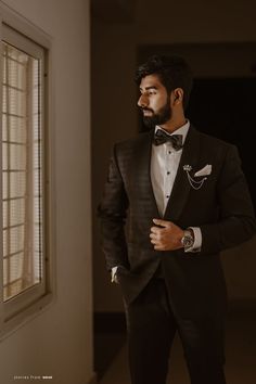 a man in a tuxedo is standing by a window with his hands on his hips