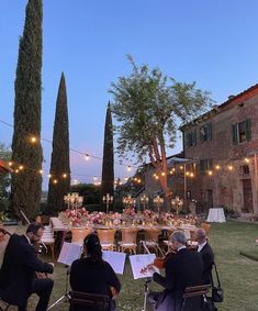 a group of people sitting around a table with musical instruments in front of them and lights hanging from the trees