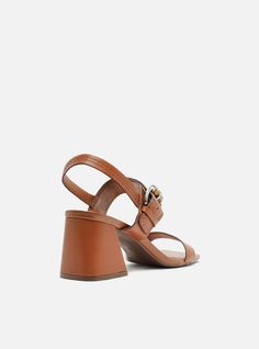 Brown sandal. The style features a high block heel and a rounded toe. It also has a mid-strap over the toes, around the heel and over the instep with a rounded metal buckle featuring a featuring a woven detail. With an insole in the same color as the sandal and the brand name engraved, it shows the foot. Why follow this trend? Step outside the box with a strappy sandal and big buckles. The Sage Mid-Block Sandal features a versatile design and comfortable fit so you can go to the office, run erra