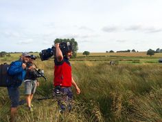 two men with camera equipment are walking through tall grass in the middle of an open field