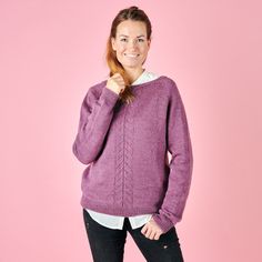 a woman wearing a purple sweater and black jeans poses in front of a pink background