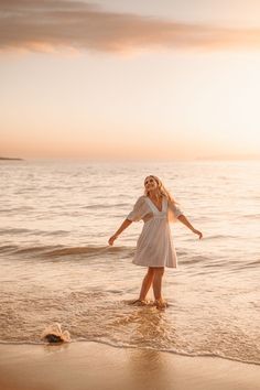 a woman in a white dress standing on the beach with her arms spread out and looking at the water