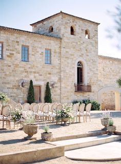 an outdoor ceremony setup with chairs and flowers in front of a stone building on a sunny day