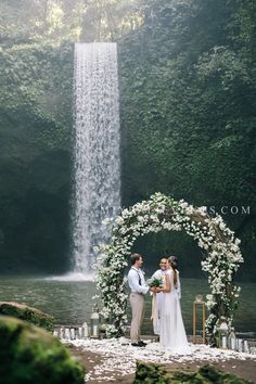 a bride and groom standing in front of a waterfall at their wedding ceremony with flowers on the altar