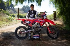 a man standing next to a red dirt bike on a dirt road with trees in the background