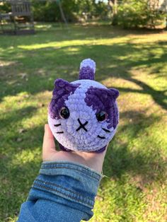 a hand holding up a small crocheted cat in the grass with trees in the background