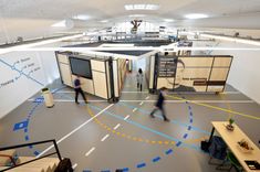 two people walking through an office building with cubicles on the floor and overhead lighting