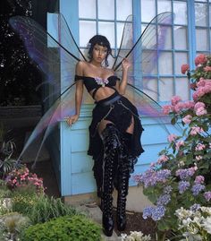 a woman dressed as a fairy standing in front of a blue building with flowers around her