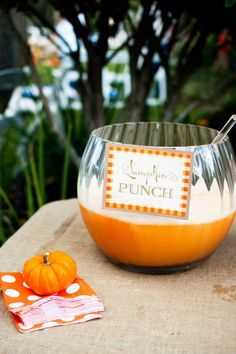 an orange colored bowl with a spoon in it on top of a wooden table next to a potted plant