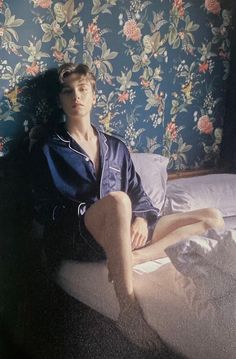 a young man sitting on top of a bed next to a wallpapered wall