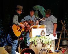 three men are playing guitars and singing in front of a barrel full of stuff on the ground