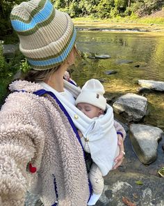 a woman holding a baby while standing next to a river with rocks and trees in the background