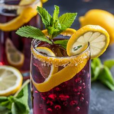 two glasses filled with red liquid and lemons on top of a gray table next to green leaves