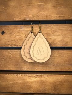 two leather tear shaped earrings hanging from hooks on wooden planks with the letter a