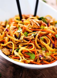 a white bowl filled with noodles and vegetables on top of a wooden table next to chopsticks