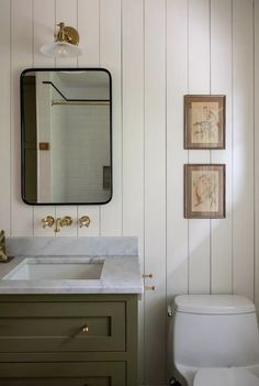 a white toilet sitting under a bathroom mirror next to a wooden sink vanity with two framed pictures on the wall