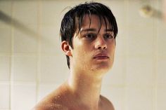 a shirtless young man standing in front of a shower head and looking at the camera