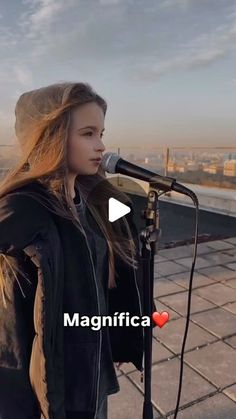 a girl standing in front of a microphone on top of a roof with the words magnifica above her