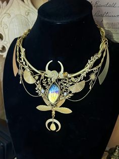 Embrace the mystique of the night sky with this enchanting Celestial Moon Goddess necklace. A true statement piece, this handcrafted treasure marries Art Nouveau elegance with ethereal fantasy elements. Key features: * Intricate gold-toned metalwork featuring delicate leaves, fans, and celestial motifs * Stunning centerpiece labradorite stone, showcasing mesmerizing flashes of blue and gold * Crescent moon shapes echoed throughout the design * Multiple layers of chains and decorative elements fo Celestial Brass Jewelry For Wedding, Celestial Brass Wedding Jewelry, Bohemian Crescent Moonstone Jewelry, Gold Fantasy Jewelry For Wedding, Gold Fantasy Wedding Jewelry, Adjustable Magical Moon Phase Jewelry, Moon-shaped Metal Jewelry For Festivals, Gold Pendant Fantasy Necklace, Handmade Moon-shaped Spiritual Necklace