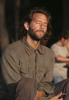 a man with long hair and beard sitting on the ground next to another man who has his eyes closed