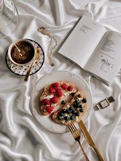 an open book and some food on a plate next to a fork, spoon and cup