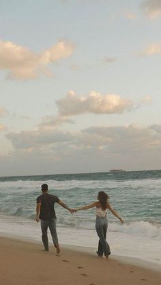 two people holding hands while walking on the beach with waves crashing in front of them