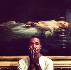 a man sitting in front of a painting with his hands covering his mouth and looking at the camera