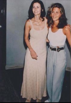 two women standing next to each other in front of a wall wearing white dresses and blue pants