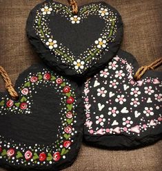 three heart shaped black stone with flowers and hearts painted on the sides, tied to twine