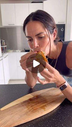 a woman is eating a slice of pizza on a cutting board in the kitchen while looking at her watch