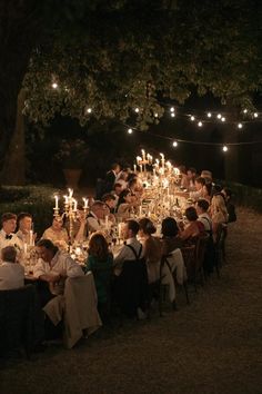 a group of people sitting around a dinner table with lit candles on it at night