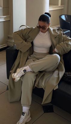 a woman sitting on top of a black couch wearing white sneakers and a trench coat